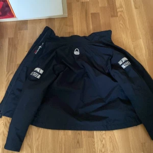 Svart vindjacka från Didriksons - Säljer en svart vindjacka från Didriksons med dragkedja framtill och reflexdetaljer på ärmarna. Jackan har huva och flera praktiska fickor. Perfekt för blåsiga dagar och utomhusaktiviteter.