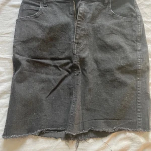 Svart jeanskjol med rå kant - Säljer en svart jeanskjol med klassisk femficksmodell och rå, fransig nederkant. Kjolen har dragkedja framtill och är i en rak, normal passform. Perfekt att styla med en enkel topp eller hoodie.