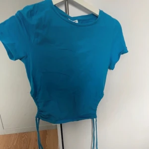 Blå croppad t-shirt med knyt - Säljer en blå croppad t-shirt med korta ärmar och knyt i sidorna. Perfekt för en avslappnad och trendig stil. Enkel att matcha med jeans eller kjol.