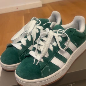 Adidas Campus gröna sneakers - Säljer ett par gröna Adidas Campus sneakers med vita ränder och vit sula. Skorna har klassisk snörning och är i mocka med extra gröna skosnören. Perfekta för dig som gillar retrostil och streetwear.
