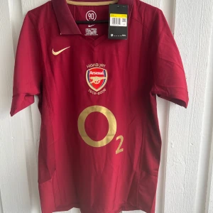 Arsenal Highbury 2005/06 fotbollströja - Säljer en klassisk Arsenal Highbury 2005/06 fotbollströja i vinrött med guldiga detaljer och O2-logga. Tröjan har korta ärmar, klubbmärke och Nike-logga framtill samt Henry 14 på ryggen. Perfekt för fans av Arsenal och Thierry Henry.