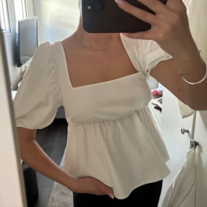Vit blus med puffärmar och öppen rygg - Säljer en söt vit blus med puffiga kortärmar och fyrkantig urringning. Blusen har en volangig nederdel och knytning i ryggen som ger en öppen rygg-detalj. Aldrig använd 