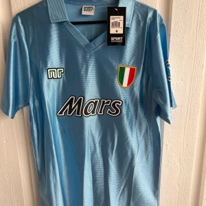 Napoli retro fotbollströja Maradona 10 - Klassisk ljusblå Napoli fotbollströja med Mars sponsortryck och klubbmärke på bröstet. På ryggen står det Maradona och nummer 10 i vitt. Tröjan har korta ärmar och krage, tillverkad i glansigt material.