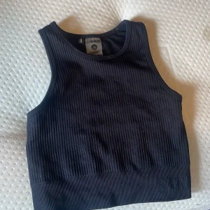 Svart ribbad linnetopp från Stronger - Säljer en svart ribbad linnetopp från Stronger. Toppen är croppad och har en tight passform med breda axelband och rund halsringning. Perfekt för träning eller som ett snyggt basplagg i garderoben.