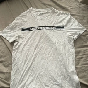 Vit t-shirt från Armani Exchange - Säljer en vit t-shirt från Armani Exchange med svart logga och text över bröstet. Klassisk rund halsringning och normal passform. Perfekt för en stilren och enkel look.