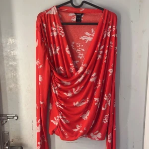 Topp  - Säljer en röd omlottopp från Lindex med vitt blommigt mönster. Toppen har lång ärm och ett snyggt draperat omlottliv. Perfekt för dig som vill ha något färgstarkt och bekvämt.