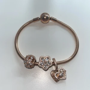 Guldfärgat armband med berlocker från Pandora - Säljer ett guldfärgat armband från Pandora med tre glittriga berlocker: två blommor och ett hjärta. Armbandet har en rund låsdel och är dekorerat med små gnistrande stenar på berlockerna.