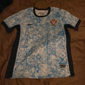 Portugal fotbollströja Nike blå/vit - Säljer en Portugal fotbollströja från Nike med unikt blått och vitt mönster, svart krage och svarta sidopaneler. Portugal-emblemet är broderat på bröstet och det står 'PORTUGAL' i nacken. Perfekt för dig som vill sticka ut på match eller träning.