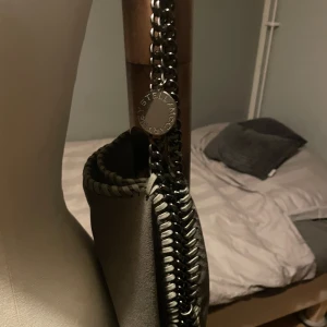 Grå axelväska från Stella  - Snygg grå axelväska från Stella med kedjehandtag och dekorativa sömmar längs kanterna. Väskan har en rund metallbricka med logotyp  som ger en lyxig känsla. Falabella modell