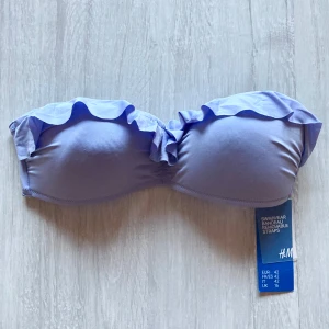 Ljuslila bandeau bikiniöverdel från H&M - Säljer en ljuslila bandeau bikiniöverdel från H&M med volangdetalj upptill. Bikinin har avtagbara axelband och en enkel, stilren design. Perfekt för sommardagar vid stranden eller poolen.