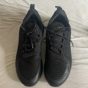 Svarta Nike Air Max 270 sneakers - Säljer ett par helsvarta Nike Air Max 270 sneakers med mesh-överdel och synlig Air Max-dämpning i hälen. Skorna har snörning och en sportig design som passar perfekt till streetwear och träning.