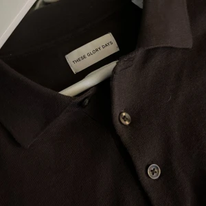 Brun pikétröja från These Glory Days - Klassisk brun pikétröja från These Glory Days i storlek M. Tröjan har lång ärm, krage och knäppning med tre knappar framtill. Perfekt för en stilren och enkel look.