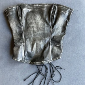 korsettopp Jaded London - Säljer en svart korsettopp från Jaded London i skinnimitation med snörning bak och dragkedja i sidan. Toppen har en cool och edgy look, perfekt för att lyfta vilken outfit som helst. Materialet är en blandning av viskos och polyuretan.