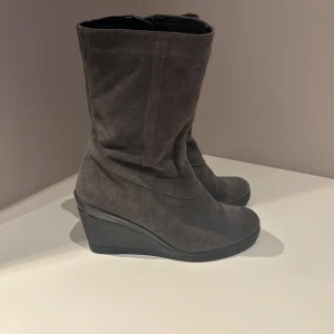 Gråa kilklack boots från Janet D. - Snygga grå boots i mocka från Janet D. med kilklack och dragkedja på sidan. Skorna har en rund tå och är perfekta för dig som vill ha både stil och komfort. Tillverkade i äkta skinn och mocka.