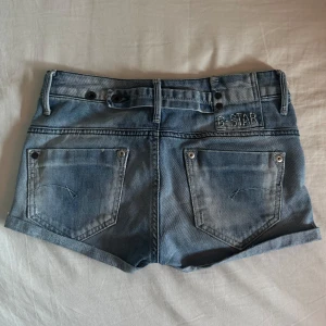 Low waisted jeansshorts från G-Star - Säljer ett par klassiska blå jeansshorts från G-Star med fem fickor, bälteshällor och uppvikta benslut. Shortsen har normal passform och är tillverkade i denim med snygga slitningar. Perfekta för sommaren!
