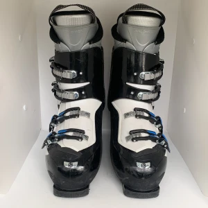 Svarta och vita pjäxor från Salomon Mission R70 - Salomon Mission R70 pjäxor i svart och vitt med fyra justerbara spännen i metall och blå detaljer. Insidan är grå och svart, och yttermaterialet är robust syntet. Perfekta för skidåkning och ger bra stöd och komfort i backen.