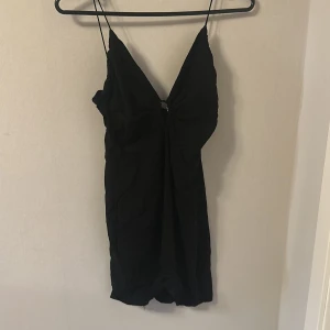 Svart kort klänning med smala axelband - Säljer en svart kort klänning från H&M med smala axelband och en snygg knutdetalj framtill. Klänningen har en figurnära passform och dragkedja i ryggen. Perfekt för dig som gillar enkel men stilren design.