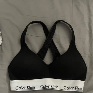 bh från Calvin Klein - Svart sport-bh från Calvin Klein med korsade axelband i ryggen och bred vit resår med logga. Bekväm och stilren design, perfekt för träning eller vardag. Mjukt material och utan byglar.