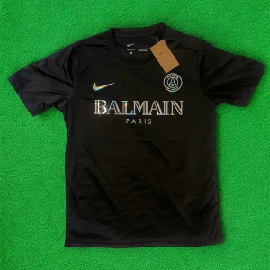 Svart PSG x Balmain fotbollströja från Nike - Säljer en svart fotbollströja från Nike i samarbete med PSG och Balmain. Tröjan har korta ärmar, rund hals och holografiska detaljer på loggor och text. Materialet är lätt och andas, perfekt för fotboll eller streetwear.