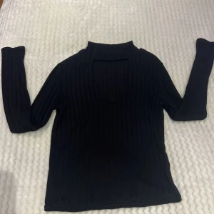 Svart ribbad polotopp med cut-out - Säljer en svart ribbad polotopp med lång ärm och snygg cut-out detalj framtill. Toppens material är stretchigt och sitter snyggt på kroppen.