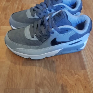 Nike Air Max 90 i grått och svart - Säljer ett par Nike Air Max sneakers i olika nyanser av grått med svarta detaljer och vit sula. Skorna har klassisk Nike-logga på sidan och AIR-text bak. Perfekta för dig som gillar sportig stil.