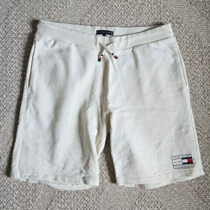 Beige shorts från Tommy Hilfiger - Snygga beigea shorts från Tommy Hilfiger med ribbad midja och snörning framtill. Klassisk logga på vänster ben och små detaljer i rött, vitt och blått på snöret. Mjukt material och en bakficka.