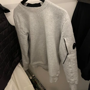 Grå sweatshirt från C.P. Company - Säljer en stilren grå sweatshirt från C.P. Company med rund halsringning och en ficka med dragkedja på ena ärmen. Klassisk passform och diskret logga på ärmen. Perfekt för en avslappnad look.