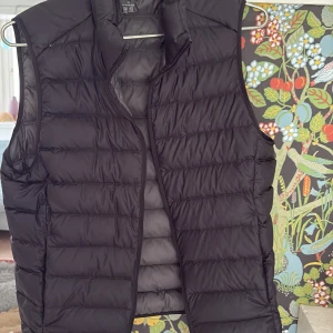 Svart dunväst från uniqlo - En stilren svart dunväst från Uniqlo med hög krage och dragkedja framtill. Västen har ett quiltat mönster och är lätt vadderad, perfekt för lager-på-lager under kyliga dagar. Passar både till jeans och träningsbyxor. Den har några små defekter men inget som märks