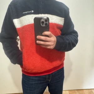 Champion fleece tröja i rött, vitt och marinblått - Säljer en mysig fleece tröja från Champion i färgerna röd, vit och marinblå. Tröjan har halv dragkedja och broderad logga på bröstet. Perfekt för kyliga dagar och har en sportig look.