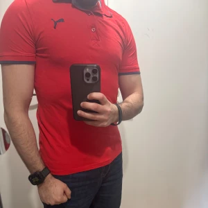 Röd piké t-shirt från Puma - Säljer en röd piké t-shirt från Puma med blå logga på bröstet. T-shirten har krage, knappar och blå detaljer vid ärmslut och krage. Perfekt för en sportig och stilren look.