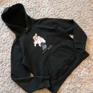Svart hoodie med Polo Bear från Polo Ralph Lauren - Svart hoodie från Polo Ralph Lauren med Polo Bear-tryck på bröstet. Tröjan har huva och magficka, samt en cool björn i skidkläder framtill. Perfekt för dig som gillar streetwear och klassiska märken.