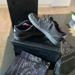 Nike Air Jordan 1 Low x Travis Scott Cactus Jack - Säljer ett par svarta Nike Air Jordan 1 Low Travis Scott Cactus Jack med omvänd swoosh och pälsdetaljer. Skorna har broderade loggor, svarta skosnören och extra röda snören. Kommer med originalkartong och bandana.