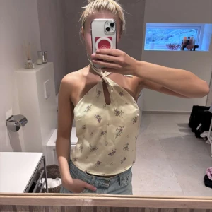 Ljusgul halterneck topp med blommigt mönster - Säljer en ljusgul halterneck topp med små blommor i lila och grönt. Toppen har knytning i nacken och baktill, samt en liten öppning framtill. Perfekt till sommaren 🌸☀️