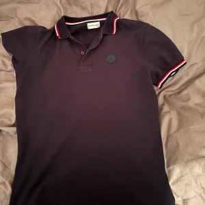 Svart piké t-shirt från Moncler - Säljer en svart piké t-shirt från Moncler med klassisk krage och knappar. T-shirten har röda och vita ränder på krage och ärmslut samt Moncler-logga på bröstet. Perfekt för en stilren look.