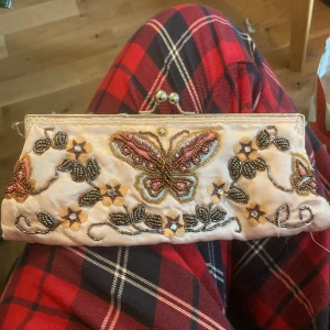 Broderad kuvertväska med fjäril och blommor - Söt kuvertväska i ljusrosa satin med broderade detaljer i form av en fjäril och blommor. Väskan har pärlor och paljetter som dekoration samt metallspänne upptill. Perfekt accessoar för att lyfta din outfit!