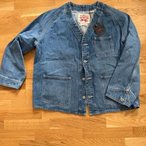 Jeansjacka med knappar från Levi's - Säljer en klassisk blå jeansjacka från Levi's med tre framfickor och knäppning framtill. Jackan har v-ringad hals och raka ärmar. Perfekt för dig som gillar en avslappnad och tidlös stil.