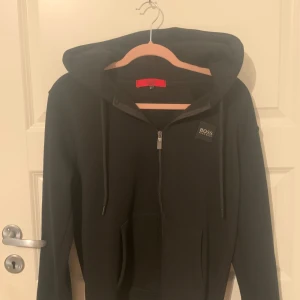 Svart hoodie från Hugo Boss - Säljer en stilren svart hoodie från Hugo Boss i storlek M. Den har en dragkedja framtill och en klassisk huva med snören. Perfekt för både vardag och avslappnade tillfällen. Märket syns tydligt på bröstet. Passar perfekt till höst och vinter!