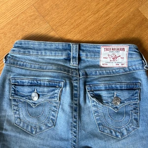True religion strl 24 - Säljer ett par ljusblå jeans från True Religion med klassiska fickor och vita sömmar. Byxorna har låg midja och bootcut, samt snygga detaljer med knappar på bakfickorna. Nyskick!❤️ modell,” Becca mid Rise bootcut flap” 