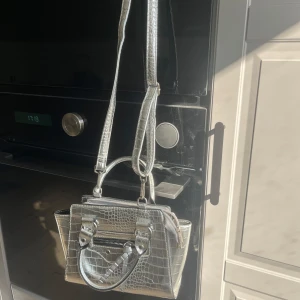 Silverfärgad axelväska med krokodilpräglat mönster - Säljer en snygg silver axelväska med krokodilpräglat mönster och justerbar axelrem. Väskan har både handtag och dragkedja framtill, perfekt för att lyfta din outfit.