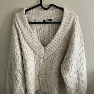 Beige stickad tröja med v-ringning från Gina Tricot - Säljer en chunky beige stickad tröja från Gina Tricot med djup v-ringning och kabelstickat mönster. Tröjan har oversized passform och långa ärmar, perfekt för lager på lager under kyliga dagar.