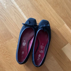 Svarta ballerinaskor med rosett från Zara - Säljer ett par klassiska svarta ballerinaskor från zara med söt rosettdetalj framtill. Skorna har rund tå, röd innersula och dekorativt blommönster under sulan. Perfekta för dig som gillar stilrena och bekväma skor. Använda men i bra skick! 