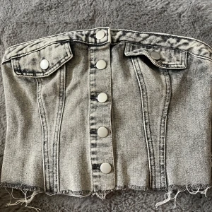 Grå jeans tubtopp med knappar - Säljer en grå tubtopp i jeansmaterial med silverfärgade knappar framtill och två dekorativa fickor upptill. Toppen har råa kanter nertill och smock baktill för bättre passform. Perfekt för en cool och trendig look.