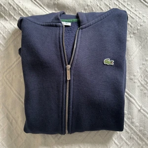 Mörkblå hoodie med dragkedja från Lacoste - Säljer en mörkblå hoodie från Lacoste med dragkedja och klassisk broderad krokodil på bröstet. Tröjan har långa ärmar, ribbade muddar och mjukt foder på insidan. Perfekt för en avslappnad och stilren look.