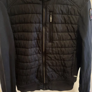 Parajumper cardigan  - Säljer en svart quiltad jacka från Parajumpers med huva och dragkedja framtill. Jackan har lång ärm, bröstficka med dragkedja och en patch på ena ärmen. Perfekt för kyliga dagar och snygg till vardags. Nypris ungefär 4 tusen kr. Strl M men passar som S