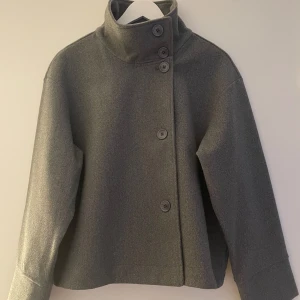 Grå kappa med hög krage och knappar - Säljer en stilren grå kappa med hög krage och stora knappar framtill. Jackan har en rak och något boxig passform med långa ärmar och kan bäras både med öppen eller stängd krage. 