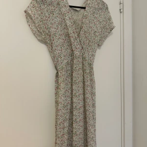 Blommig omlottklänning med knytband - Söt långklänning med blommigt mönster i rosa och grönt på vit botten. Klänningen har korta ärmar, omlottmodell och knytband i midjan för en fin passform. Perfekt för sommarens alla tillfällen.