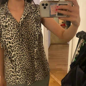 Leopardmönstrad blus med volang - Säljer en snygg blus med leopardmönster och volangdetaljer längs framsidan. Blusen har krage och knäppning framtill, är ärmlös och i ett lätt material. Pris kan diskuteras