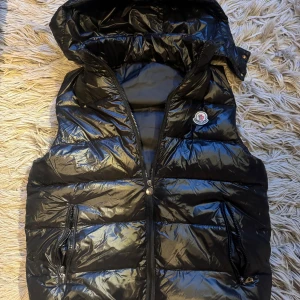 Moncler väst - Säljer en svart glansig dunväst från Moncler med huva och dragkedja framtill. Västen har två fickor med dragkedja och Moncler-logga på bröstet. Perfekt nu till våren.10/10 skick och inga defekter! Moncler påse kommer med. Bara att höra av sig vid minsta fundering!