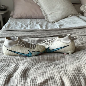 Vita Nike Mercurial Air Zoom fotbollsskor - Säljer ett par vita Nike Mercurial Air Zoom fotbollsskor med blått och svart mönstrad swoosh och rosa detaljer på sulan. Skorna har snörning och är designade för att ge bra grepp på planen. Perfekta för dig som vill sticka ut på matchen!