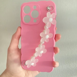 Preppy rosa iPhone 14 Pro mobilskal  💕 - Säljer ett gulligt rosa mobilskal med vågig kant, dekorerat med vita blommor och pärlor på ett band. Perfekt accessoar för att göra mobilen extra söt och personlig. HELT OANVÄND 💞